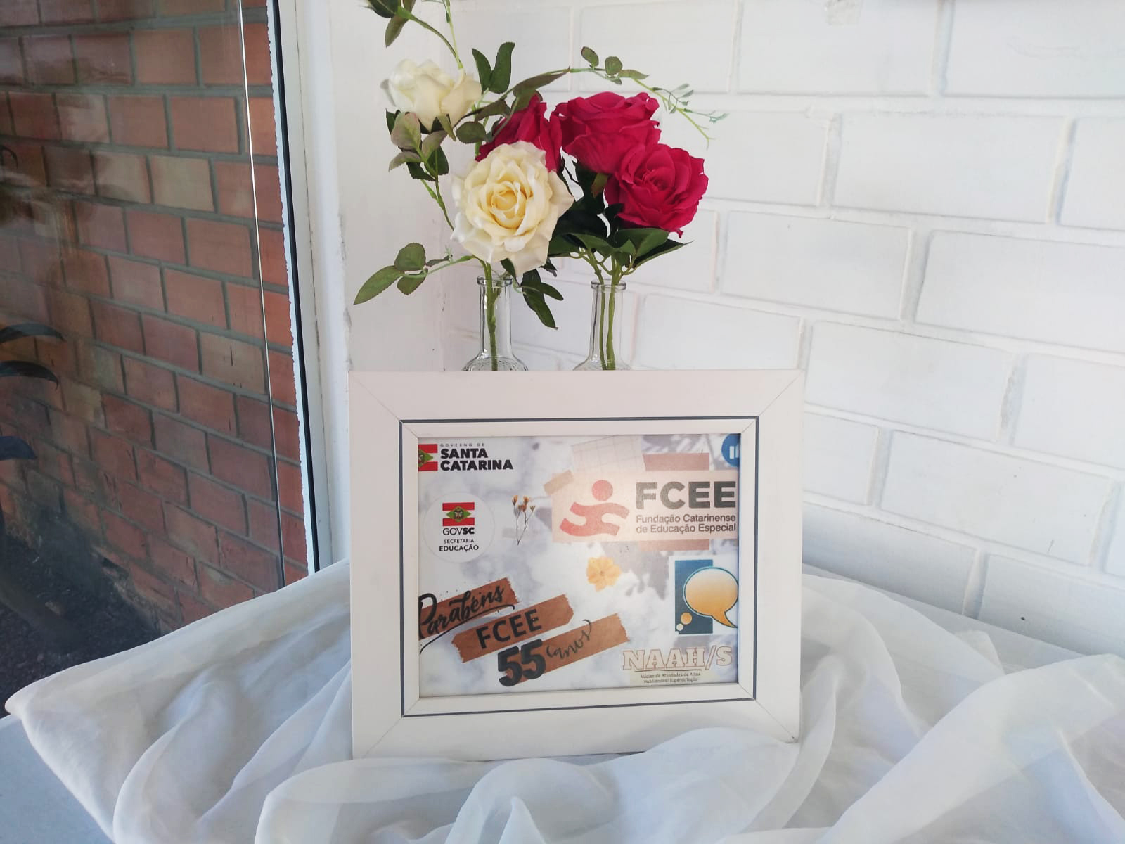 Vaso de flor com rosa vermelhas e brancas sobre mesa com toalha branca e cartaz escrito Parabéns FCEE 55 anos