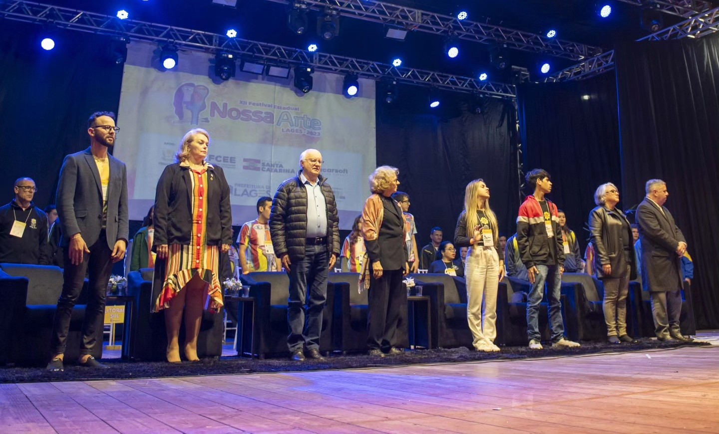 Oito pessoas em pé em um palco. Quatro mulheres e quatro homens todos brancos em posição de sentindo durante execução do hino nacional na abertura do Festival Nossa Arte em Lages. Usam casacos e blazer nas cores preta e azul marinho. Atrás há um telão com a frase Festival Nossa Arte 