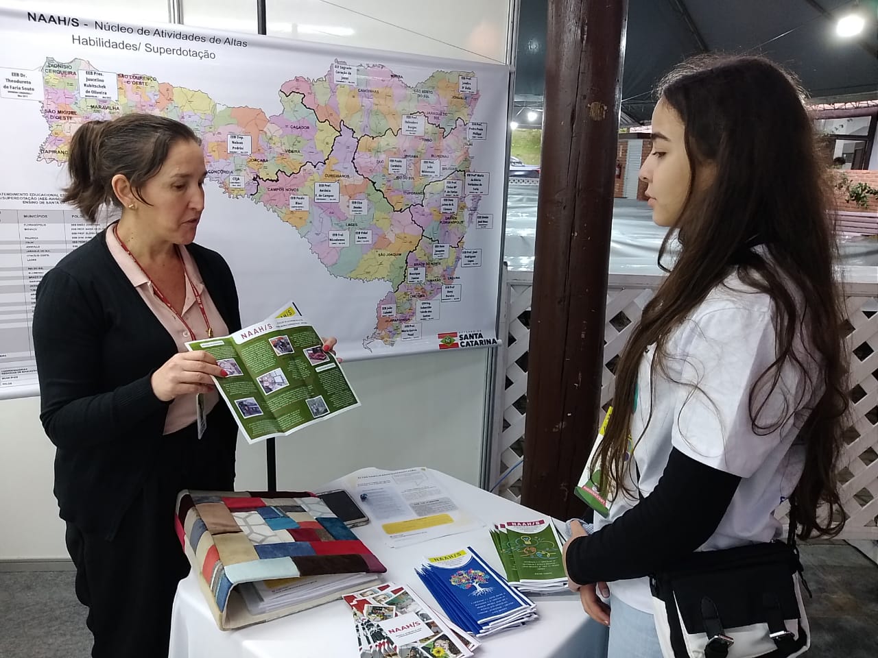 Uma mulher branca, cabelos claros com blazer preto, mostra uma folder da cor verde para uma moça branca, cabelos pretos longos, trajando camisa branca. As duas estão ao redor de uma mesa com vários folderes. Atrás das duas pessoas há um mapa de Santa Catarina