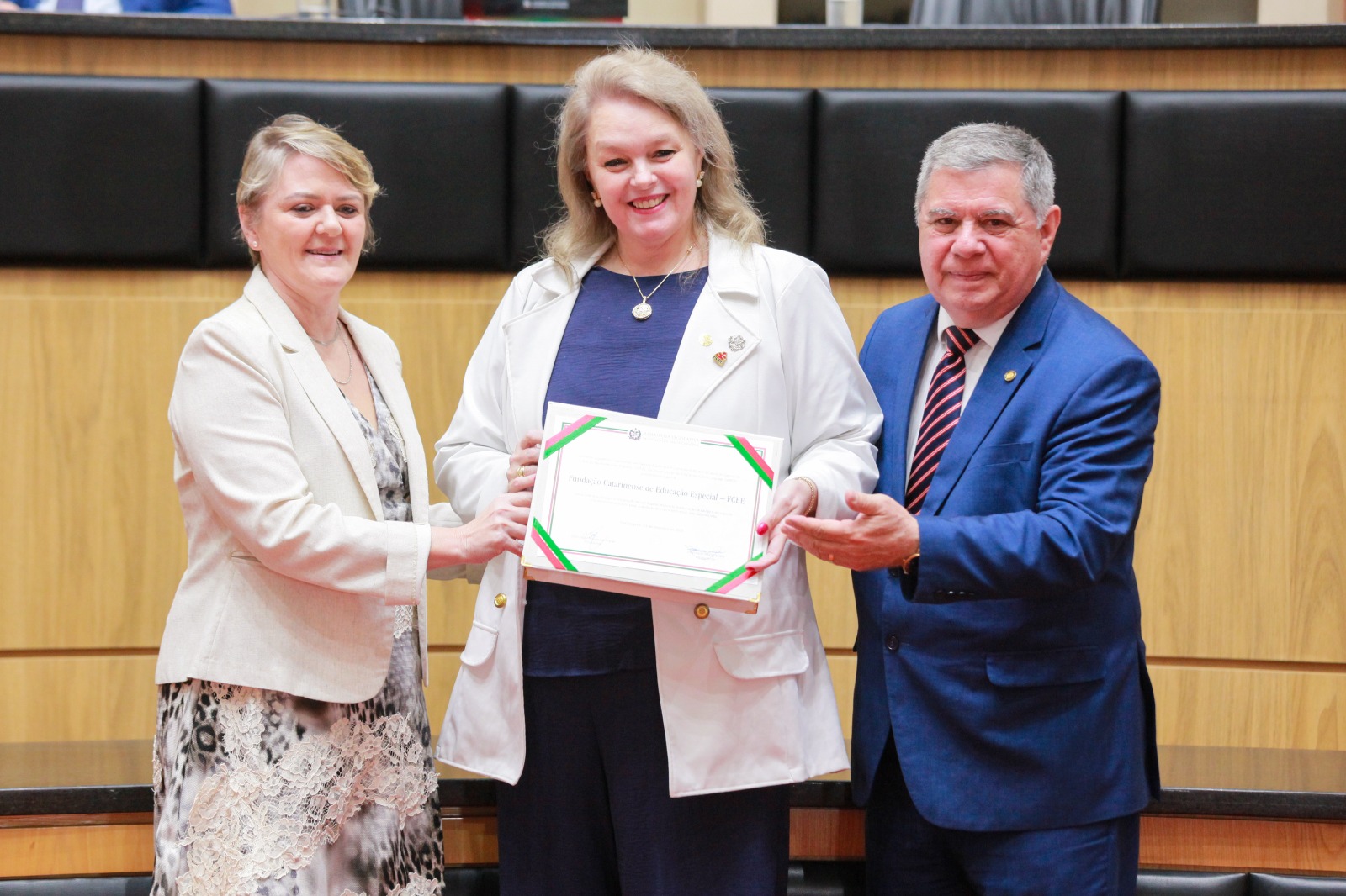 Três pessoas posam para foto com um certificado. As duas primeiras mulheres brancas trajam bleizer branco, último, um homem, branco, está de paletó azul marinho. todos seguram o certificado