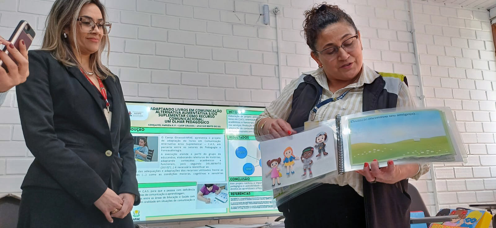 duas mulheres em sala de aula, uma delas segura pasta com desenhos coloridos