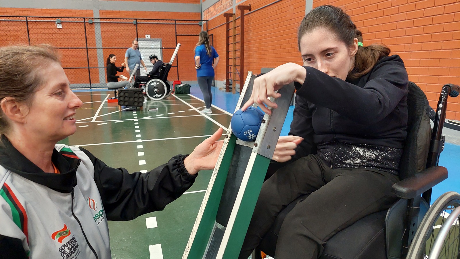 A imagem mostra uma atleta em cadeira de rodas em um ginásio, vestindo agasalho preto, sendo auxiliada por sua treinadora, que veste agasalho branco. A atleta disputa uma partida de bocha paralimpica. Na imagem ela esta empurrando uma bola azul, que desliza sobre uma calha de madeira da cor verde