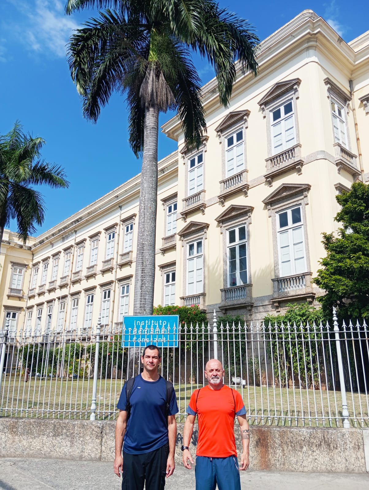 2 homens posam para foto em frente a prédio histórico, céu azul ao fundo