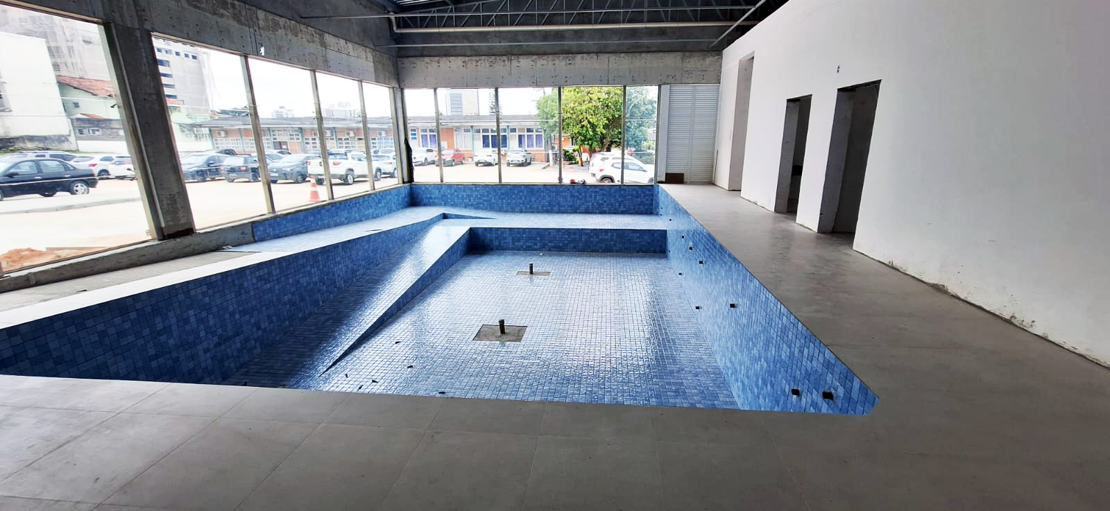 Piscina vazia em prédio em obras, coberta e com janelas ao fundo. 