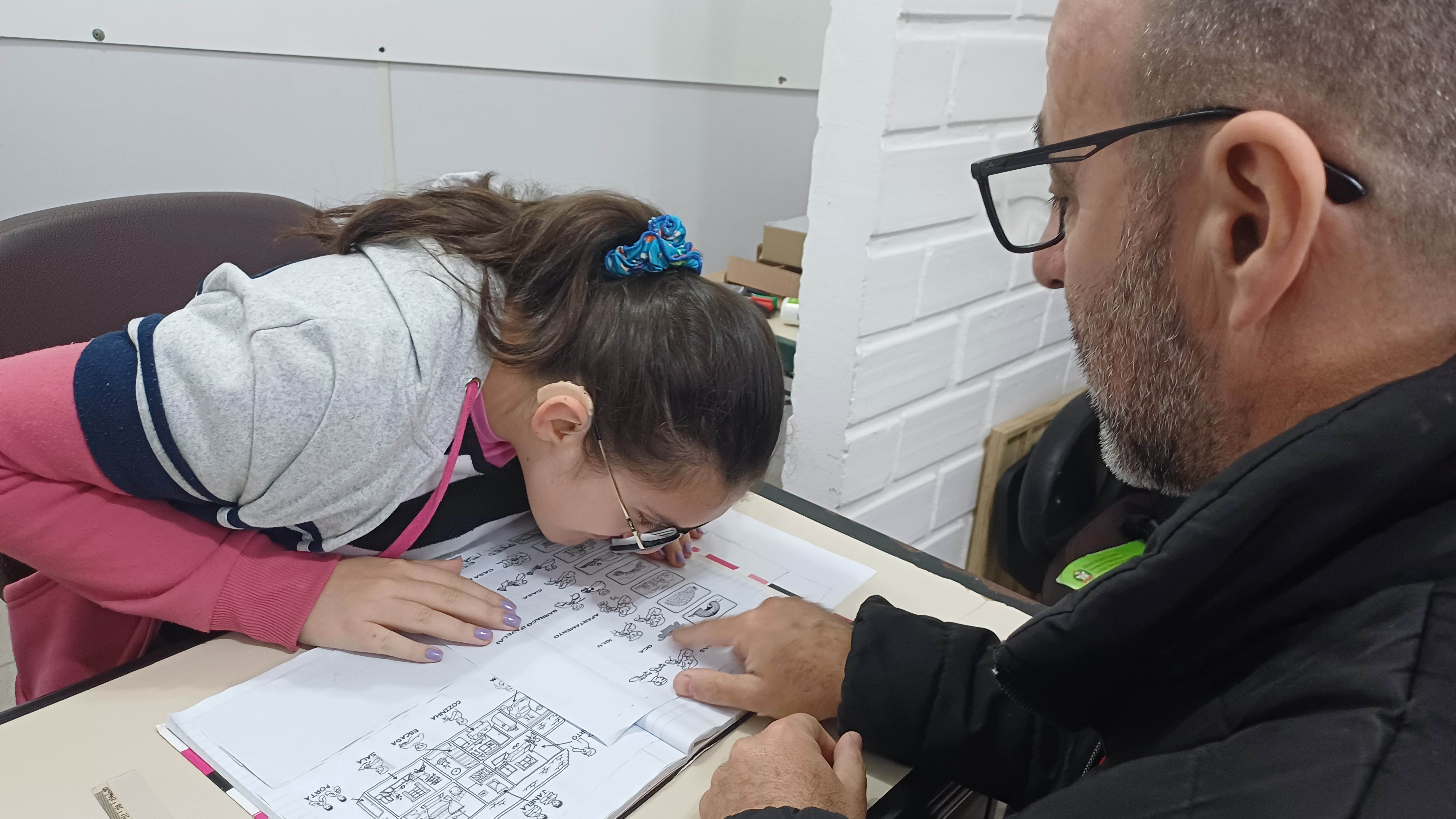 Duas pessoas sentadas em mesa de estudo, um professor e uma menina