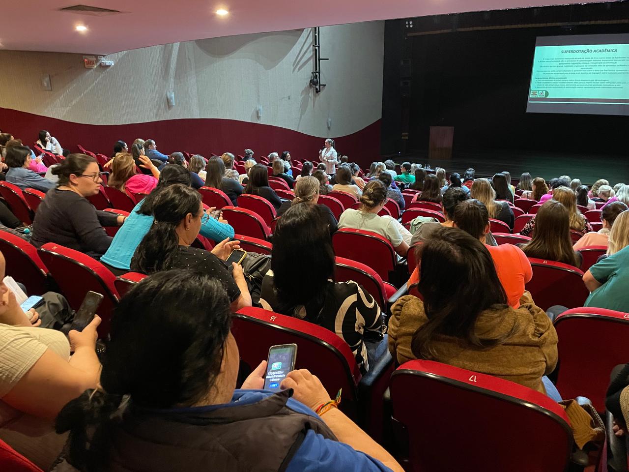 dezenas de pessoas sentadas em auditório, ao fundo palco. 