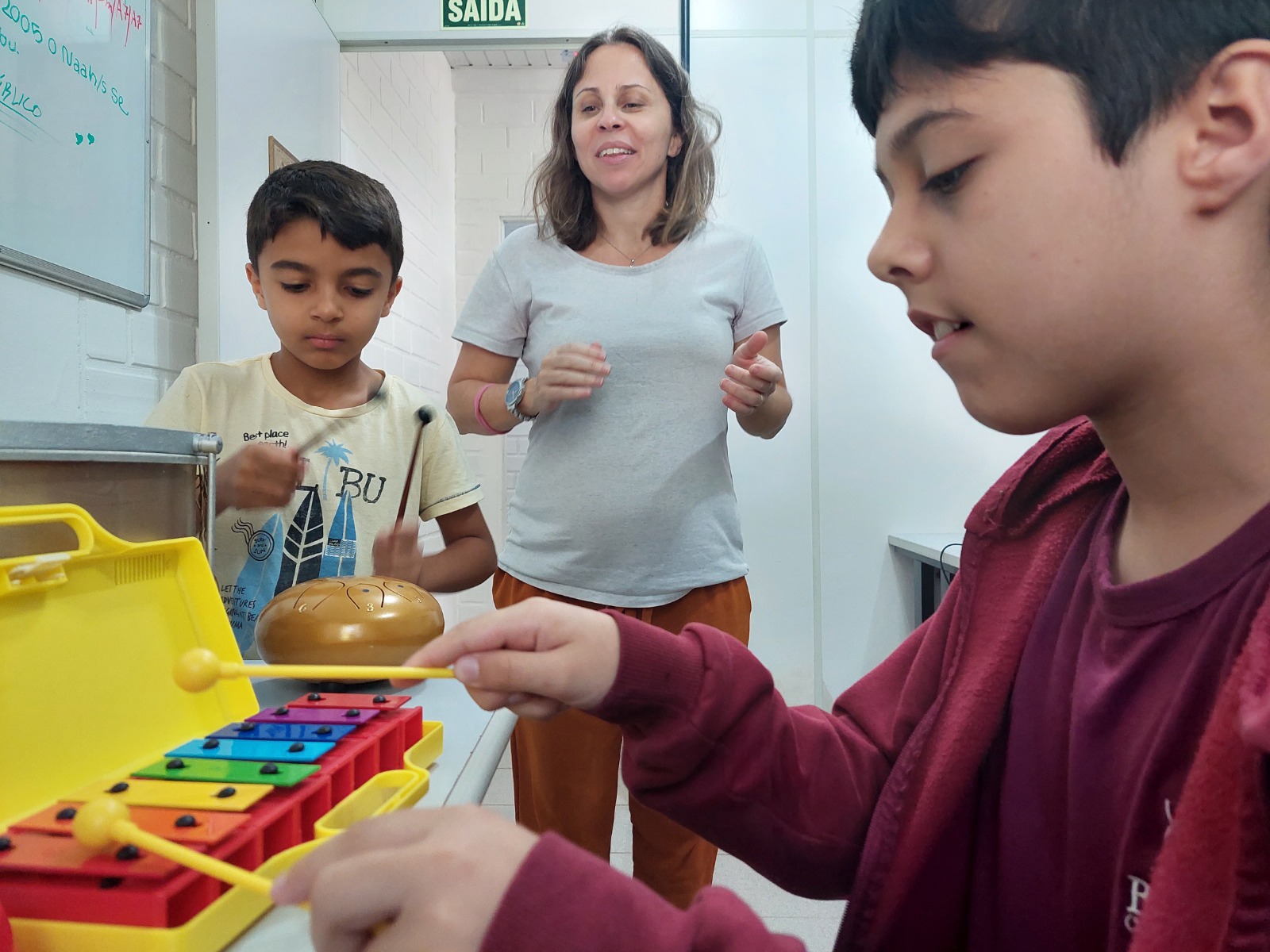 A imagem mostra três pessoas. À direita um adolescente moreno toca uma miniatura em brinquedo de um piano amarelo. O garoto é moreno e usa moleton marrom. Ao centro uma mulher branca, cabelos loiros, grávida, observa. Ela sorri. Do seu lado há um menino, moreno, que observa a cena.