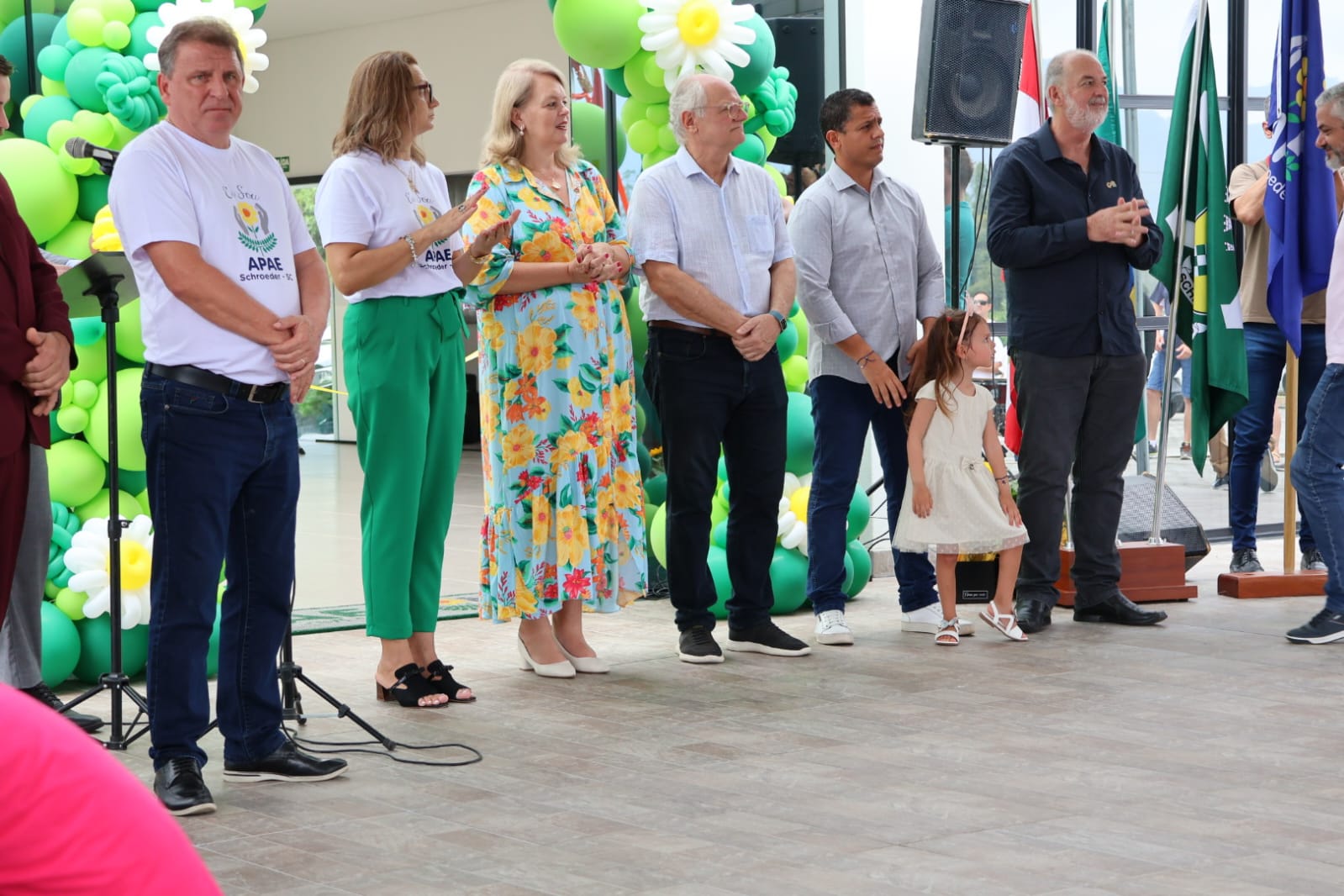 A imagem mostra seis pessoas em pé em ambiente coberto. São duas mulheres e quatro homens. Elas são autoridades e estão partipando da cerimônia de inauguração do prédio da Apae do município de Schroeder. Entre as autoridades está uma criança. Uma menina entre 3 a 5 anos de idade, que traja vestido amarelo claro. Atrás das pessoas aparecem balões nas cores verde e amarelo, que fazem parte da decoração, e duas bandeiras, uma verde, do município, e outra azul, que não está bem visível no que está estampada nela.