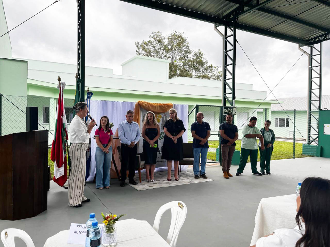 Nove pessoas em pé em cerimônia em quadra coberta, uma mulher segura um microfone, ao lado um porta-bandeiras, ao fundo grama e prédio térreo na cor verde água. 