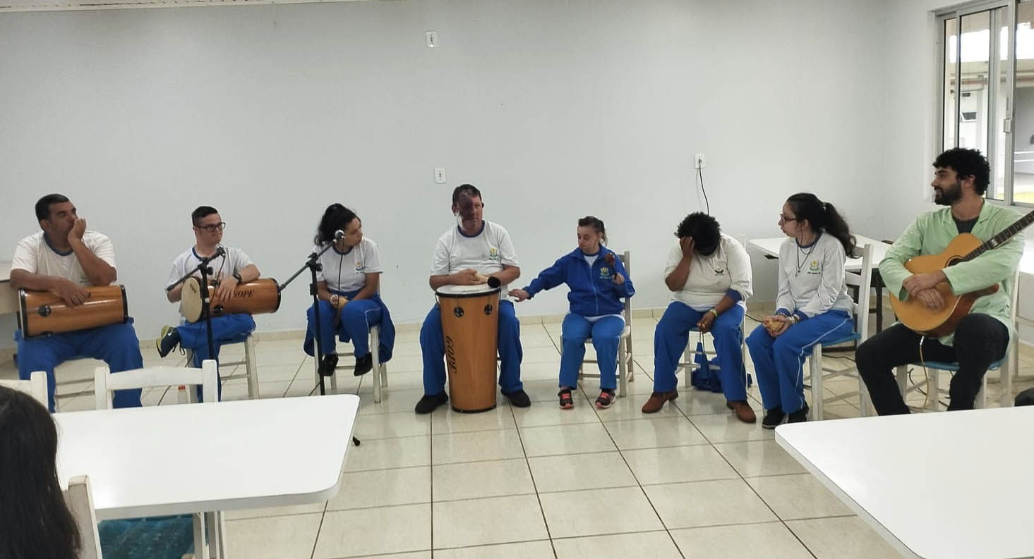 Ambiente interno, sala de aula, oito pessoas sentadas fazem apresentação de música, seguram tambores, violão e outros instrumentos, microfones com pedestal. Sete deles usma uniforme calça azul e camiseta branca. 