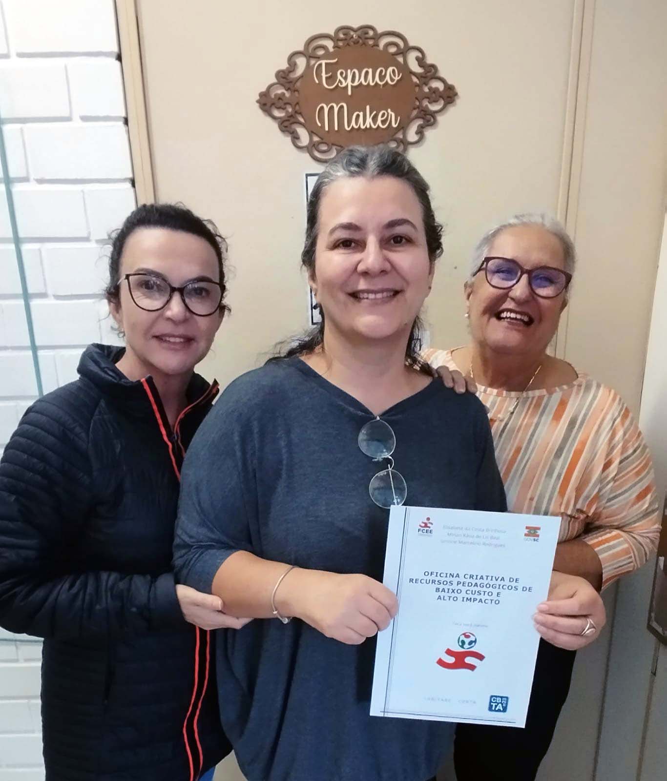 3 mulheres sorrindo segurando livro posam para foto, ao fundo placa escrito Espaço Maker