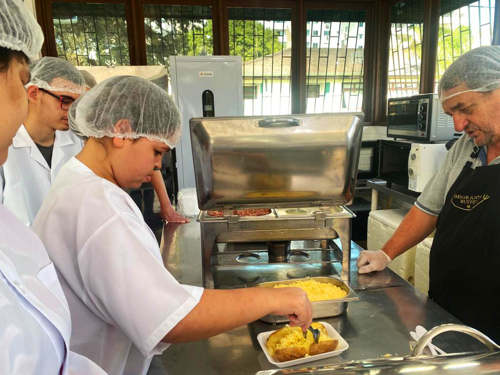 cinco pessoas com toucas e avental de gastronomia, menina segura prato de comida, professor orienta, cozinha industrial. 