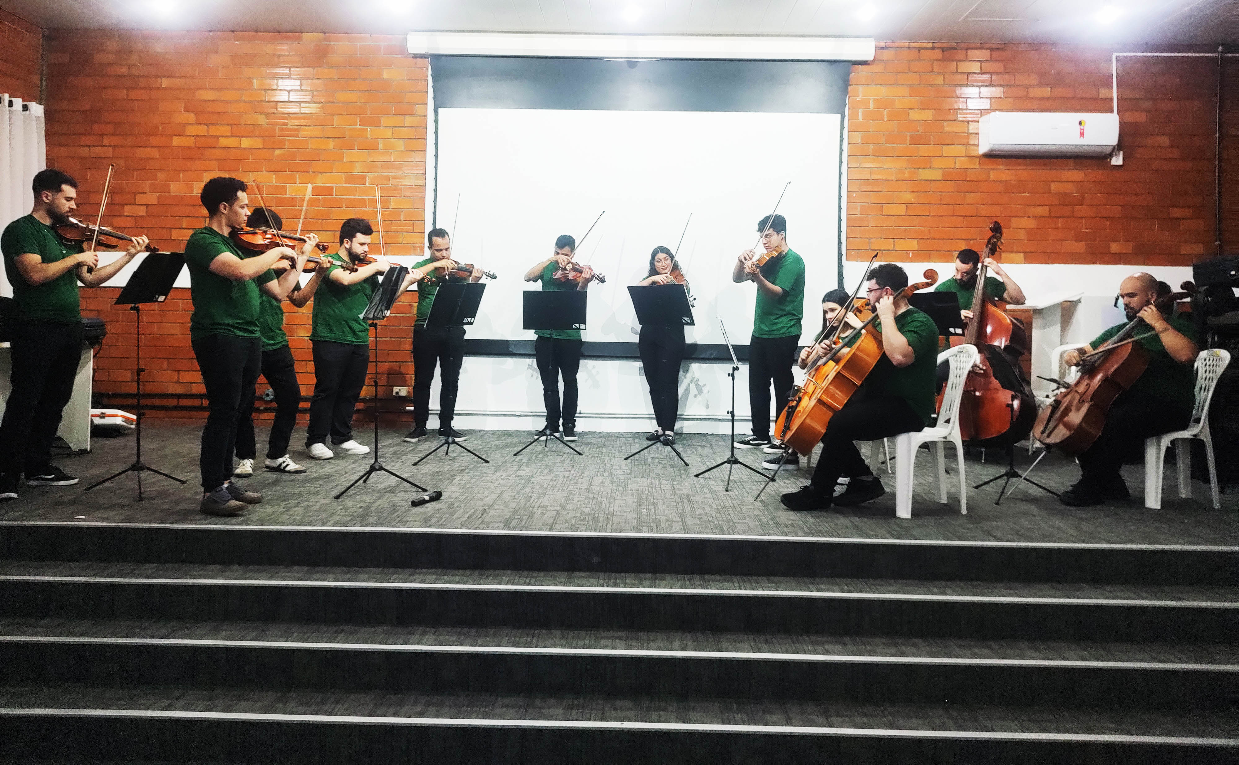 10 pessoas em palco, com instrumentos como violino, viola, contrabaixo, baixo, camiseta verde, ambiente interno