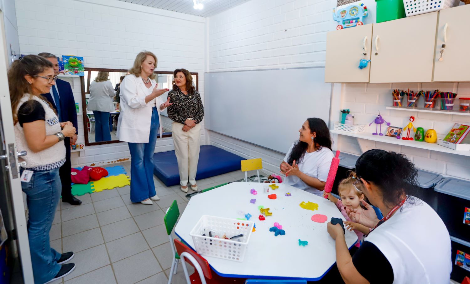 A imagem mostra duas mulheres ao centro de uma sala em pé. Uma é loira, veste blaze branco e calça jeans a outra é morena, cabelos pretos, com blusa preta e calça beje. A mulher loira está com mão estendida a explicar algo. A sua frente há duas mulheres sentadas ao redor de uma mesa com uma criança. sobrea mesa há pequebas peças de brinquedos coloridos As das mulheres são morenas e ventem jalecos brancos. À esquerda uma mulher e homem, em pé, ambos morenos, observam a cena. A mulher é morena, de óculos e jaleco cinza, o homem moreno está de paletó azul marinho.