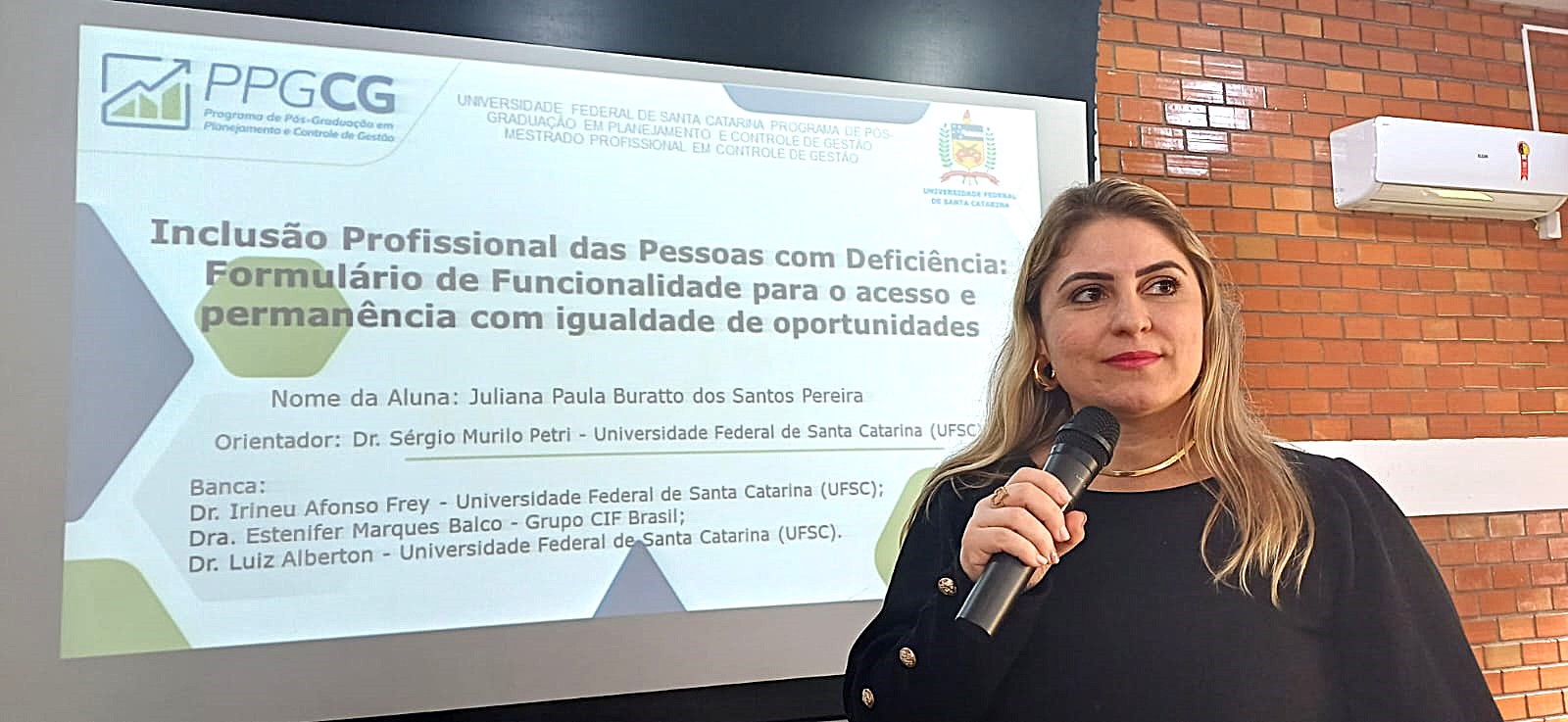 Uma mulher branca , loira, vestindo uma blusa preta está em pé, segurando um microfone. Atrás dela surge uma imagem no retroprojetor com os dizeres: inclusão profissional das pessoas com deficiência: formulários de funcionalidades para o acesso e permanência com igualdade de oportunidades 