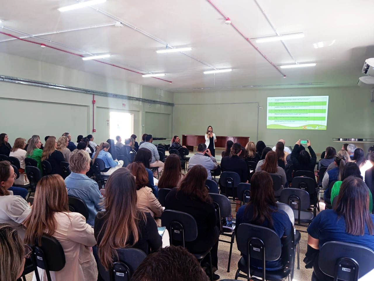 pessoas sentadas em auditorio, ao fundo pessoa em pé falando em microfone