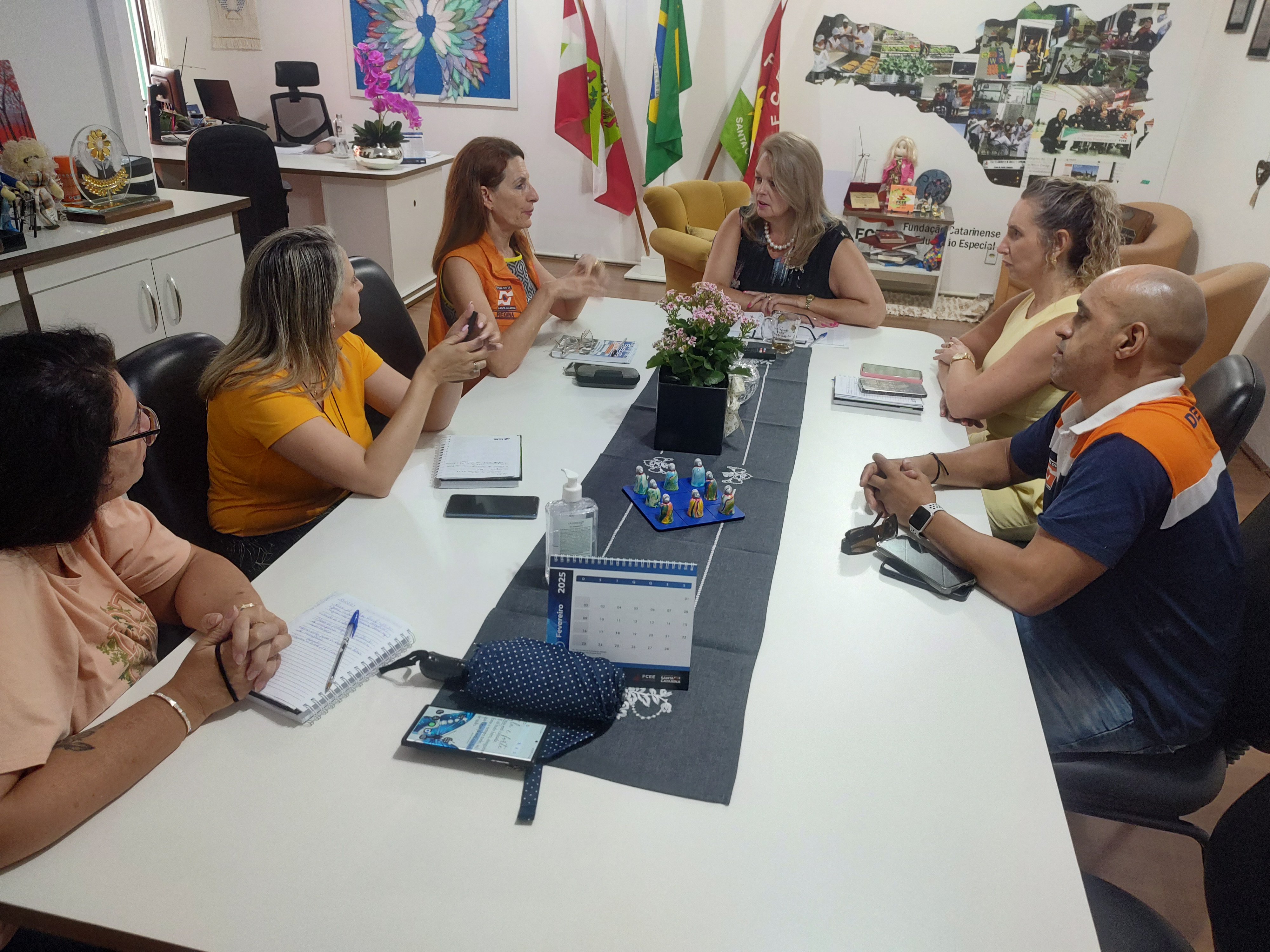 seis pessoas sentadas em mesa de reunião, coletes laranjas da defesa civil