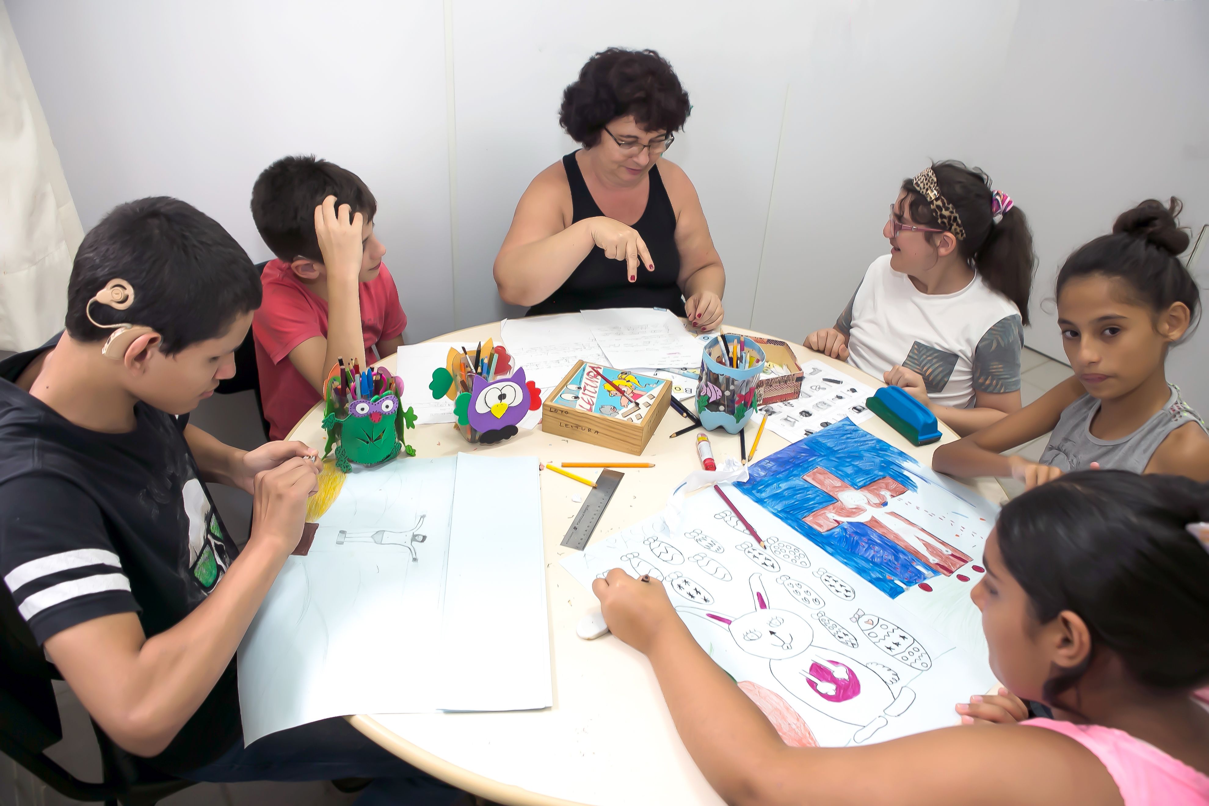 Grupo de estudantes sentados em mesa redonda com professora fazendo sinais. 