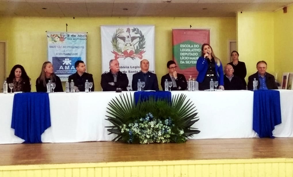 Gerente da FCEE Juliana Buratto fala ao microfone em pé no palco junto com a mesa de autoridades na abertura do evento