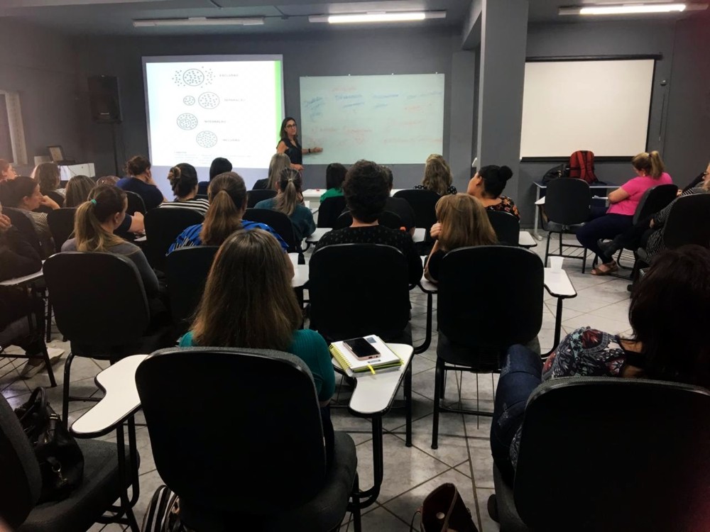 Imagem de sala de aula lotada com professora em frente a telão de projeção