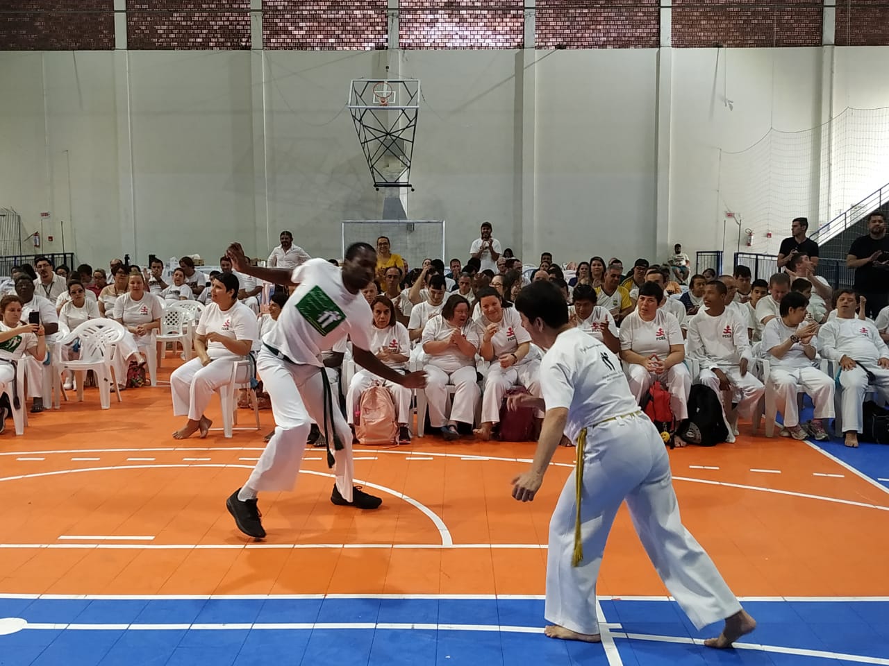 Dois capoeiristas vestidos de branco jogam diante de público sentado ao fundo em ginásio. Todos vestem calças e camisetas brancas. 