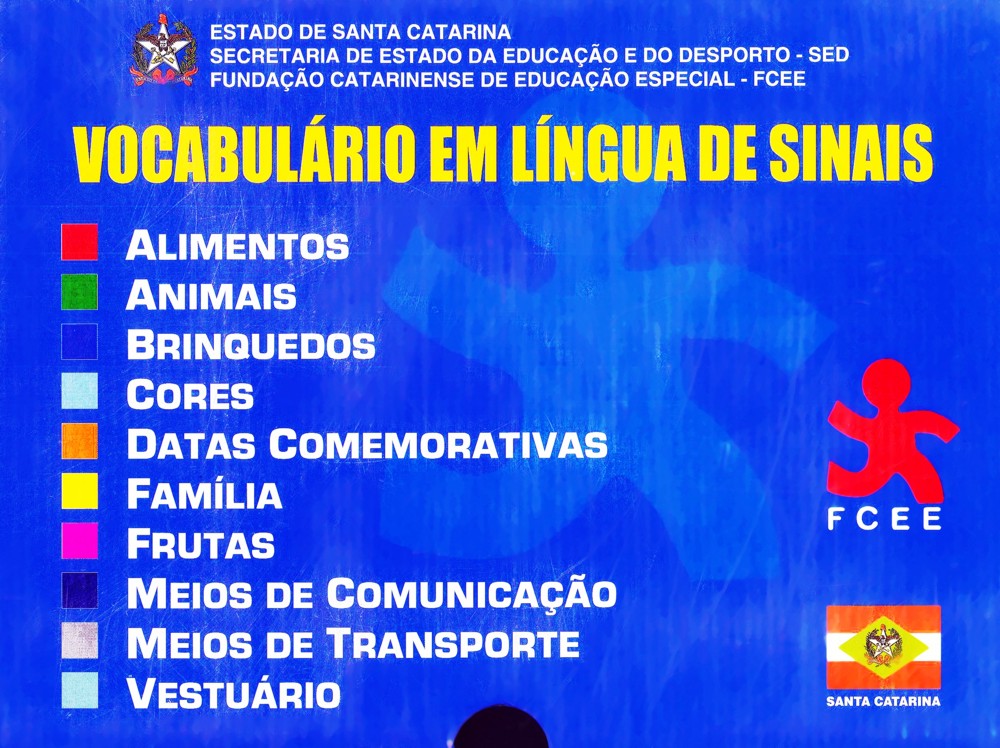 Capa da publicação com o título "Vocabulário em Língua de Sinais" e os subtítulos dos temas abordados: Alimentos, Animais, Brinquedos, Cores, Datas Comemorativas, Família, Frutas, Meios de Comunicação, Meios de Transporte e Vestuário. 