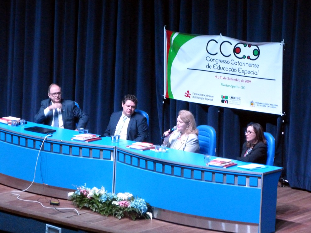 Vista do palco do auditório, com quatro pessoas sentadas na mesa de autoridades 