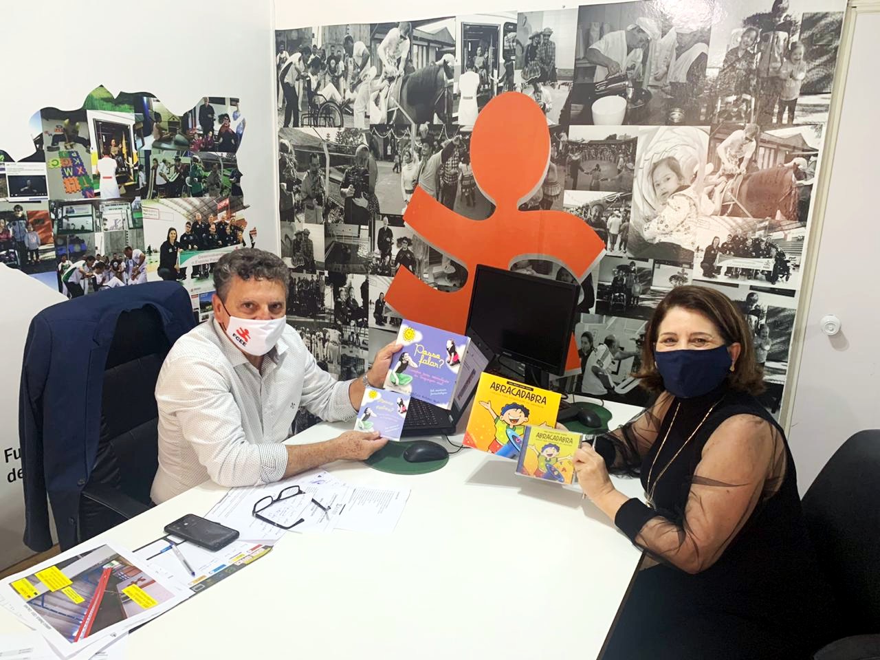 Um homem e uma mulher sentados em escritório, na mesa alguns livros. 