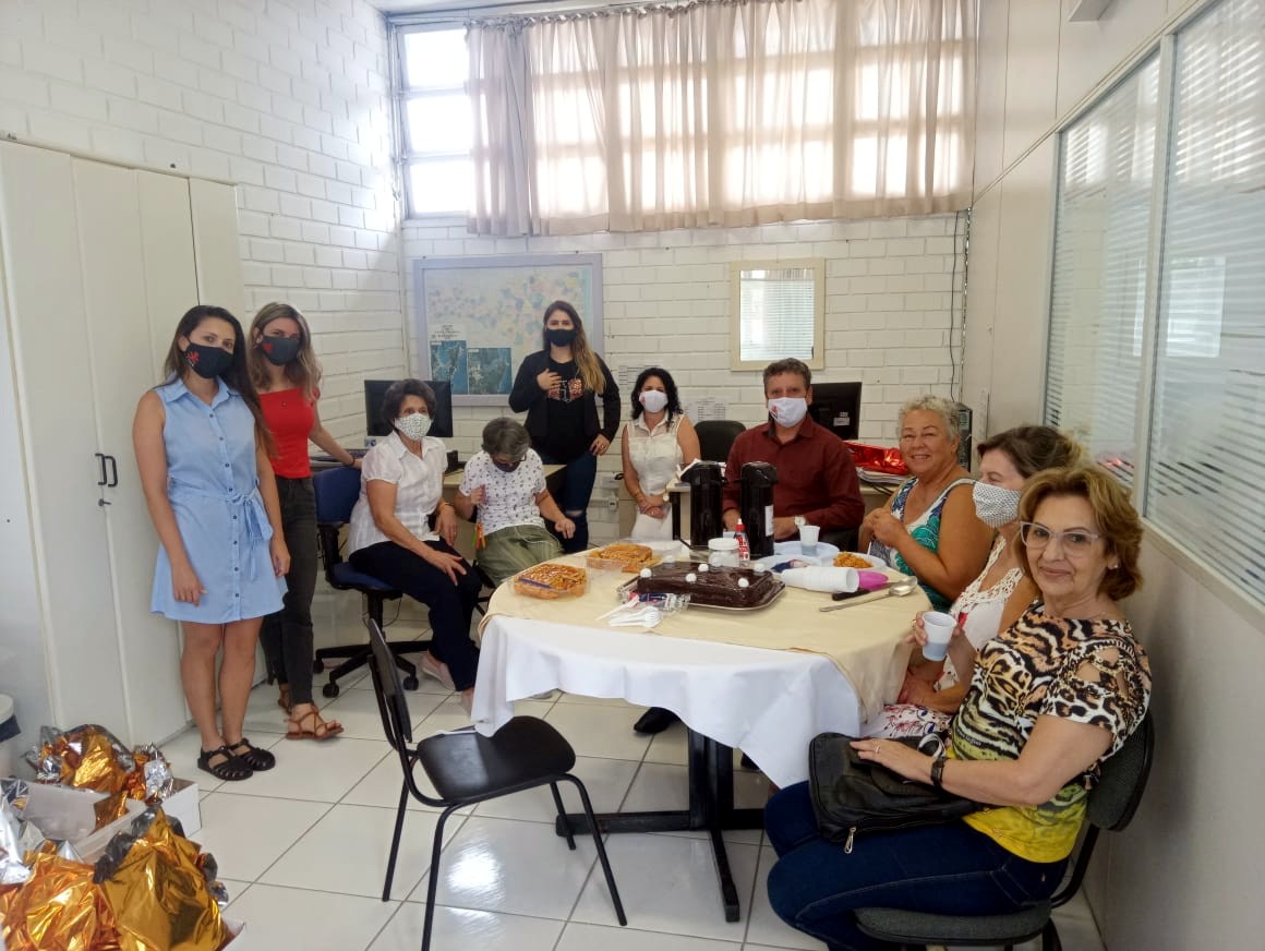 Grupo de mulheres sentadas em torno de mesa, ao centro um homem de camisa vermelha. 