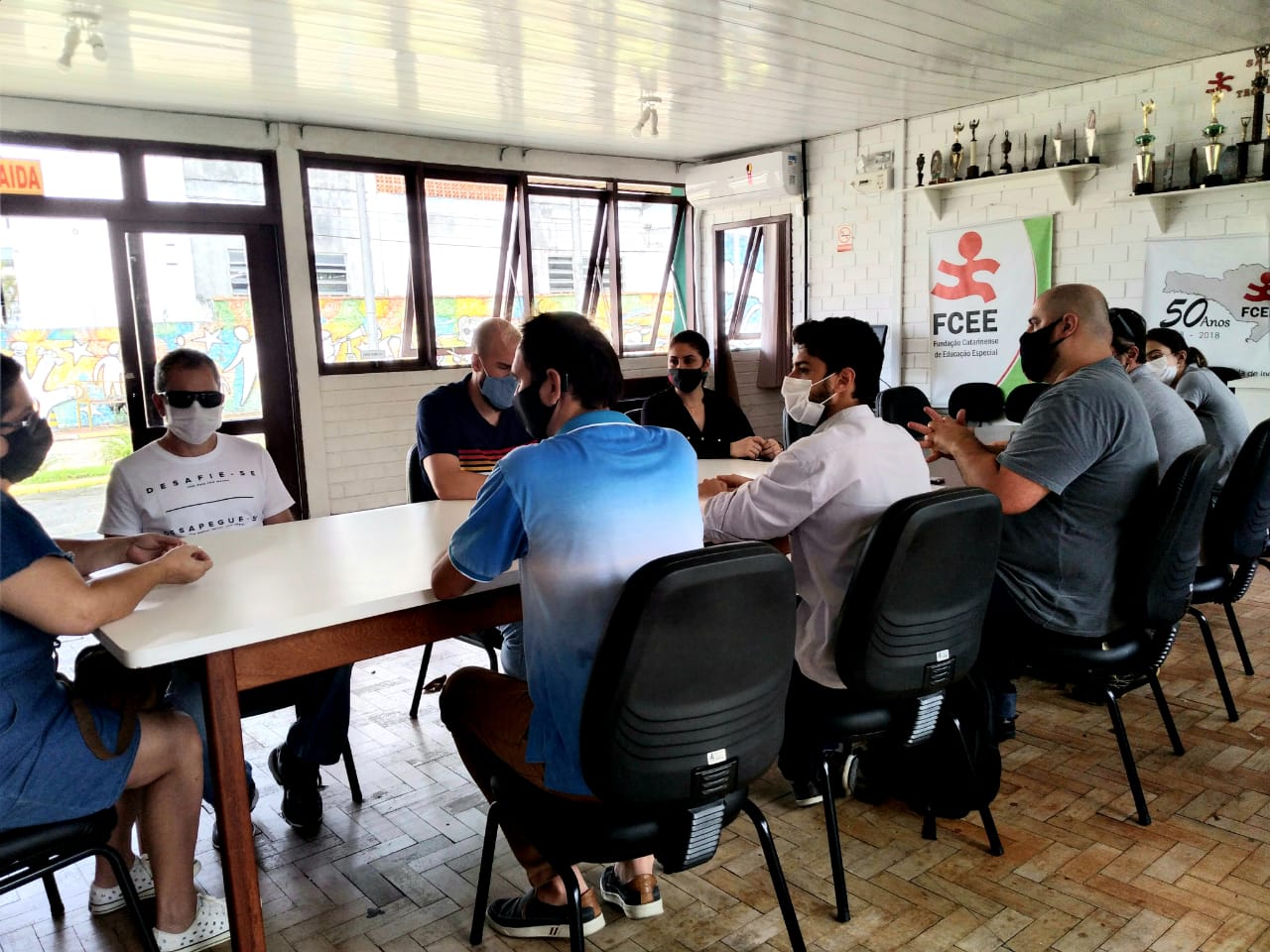 Dez pessoas sentadas em mesa de reunião, sala com janelas abertas, uso de máscaras, homens e mulheres. 