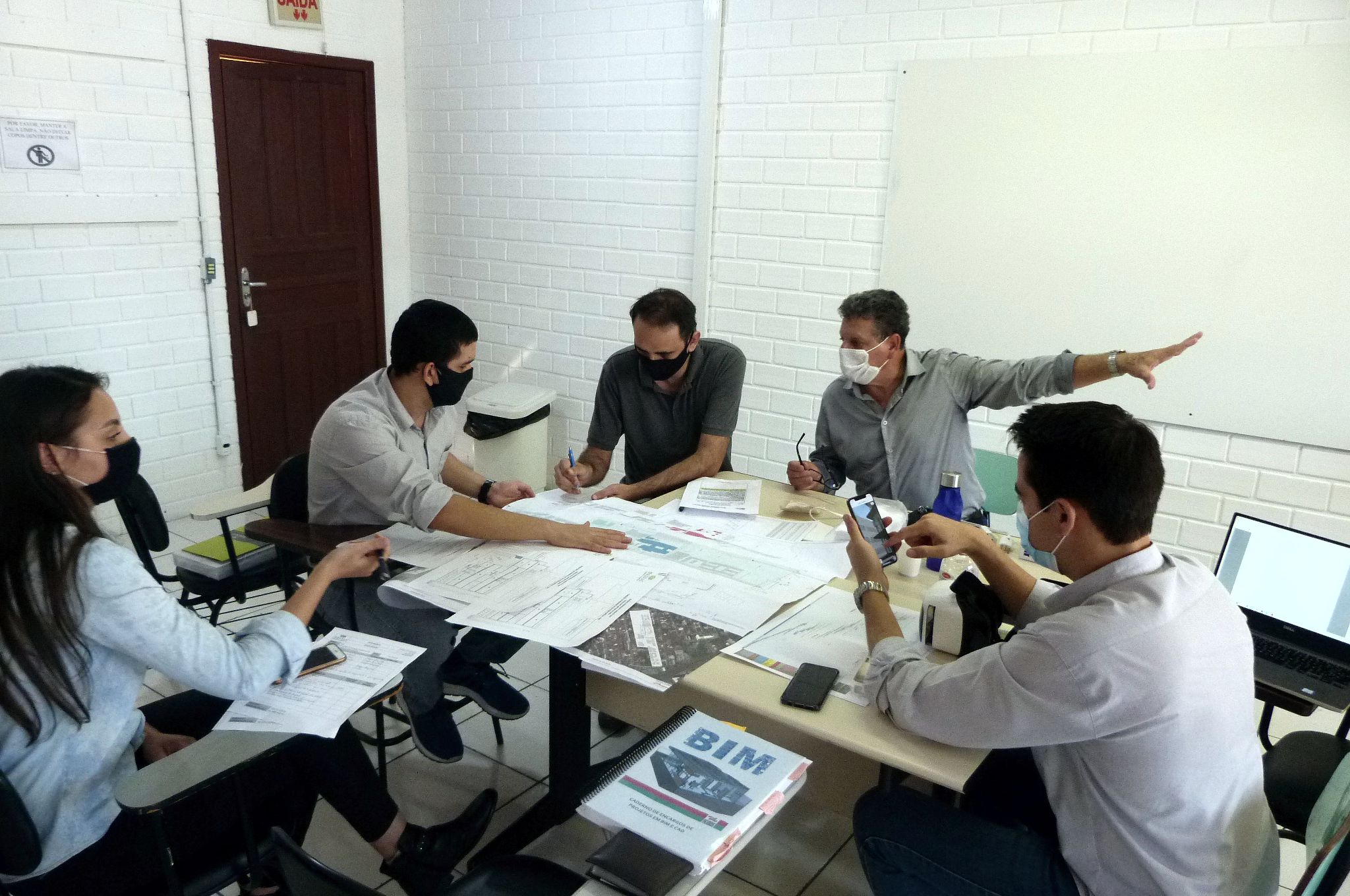 Cinco pessoas sentadas em volta de uma mesa dentro uma sala, uma mulher e quatro homens, discutindo e olhando para folhas grandes de papel sobre a mesa. 