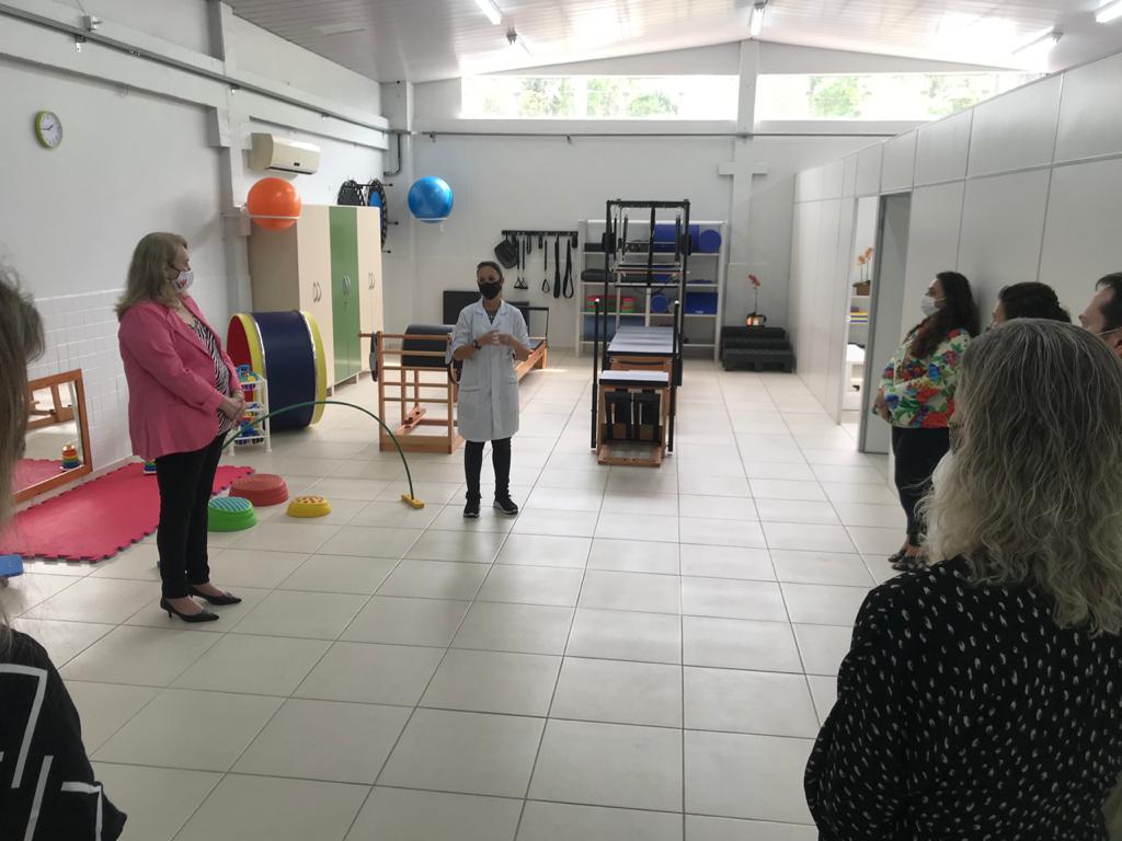 Cerca de 10 pessoas em pé, em uma sala ampla, ao centro uma mulher com jaleco branco fala, ao fundo aparelhos de reabilitação, bolas de pilates e outros materiais. 