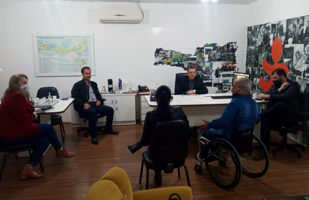 Seis pessoas sentadas em sala interna, um homem cadeirante com cabelos brancos