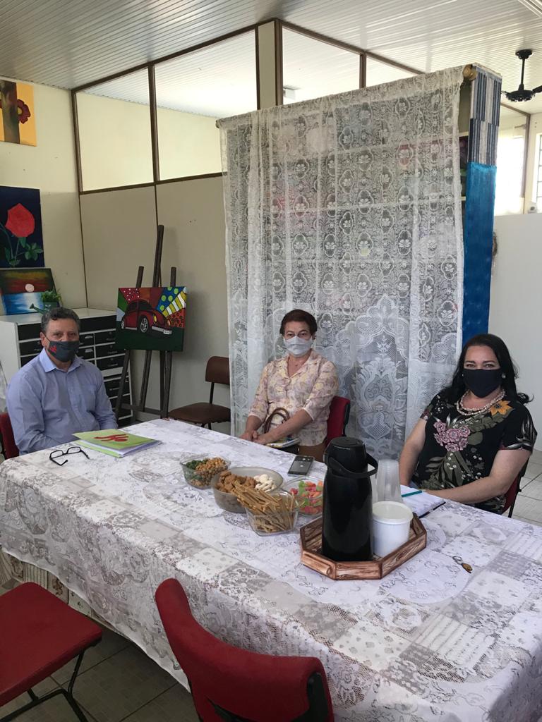 Quatro pessoas sentadas em ambiente interno, mesa com toalha crochet, 1 homem e 3 mulheres