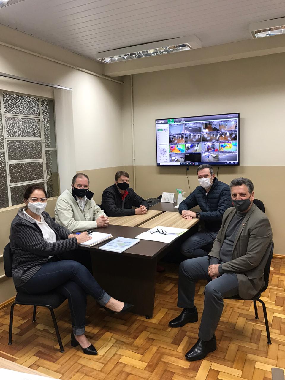 Cinco pessoas sentadas em mesa de reunião dentro de sala. Uma mulher e 4 homens. 