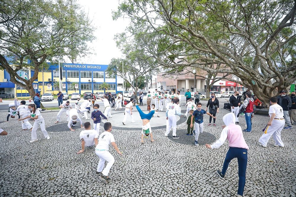 Grupo de crianças jogando capoeira, praça, árvores, ambiente aberto.