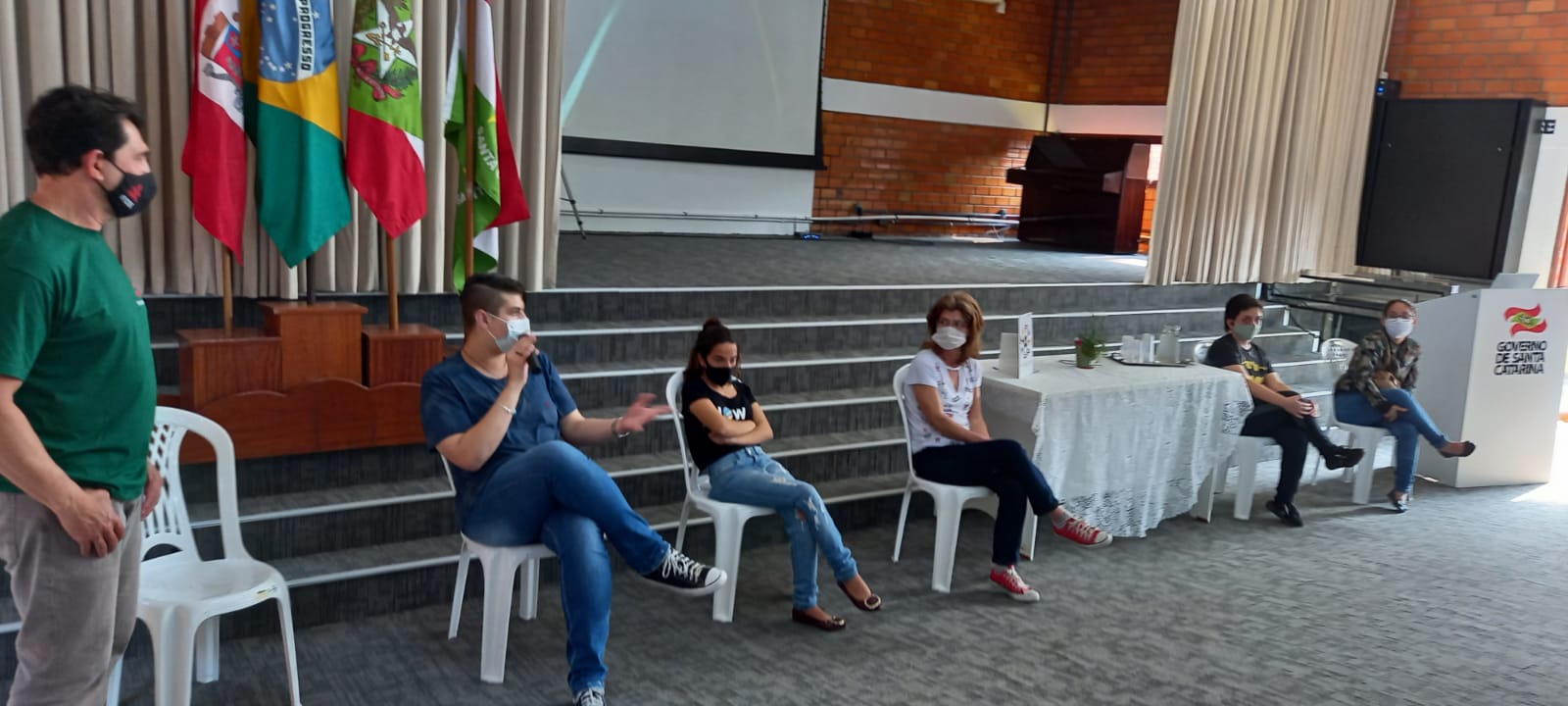 Pessoas sentadas, ambiente interno, palco ao fundo, bandeiras