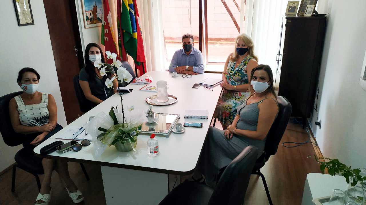 Cinco pessoas sentadas em mesa de reunião dentro de sala. 4 mulheres e 1 homem. 