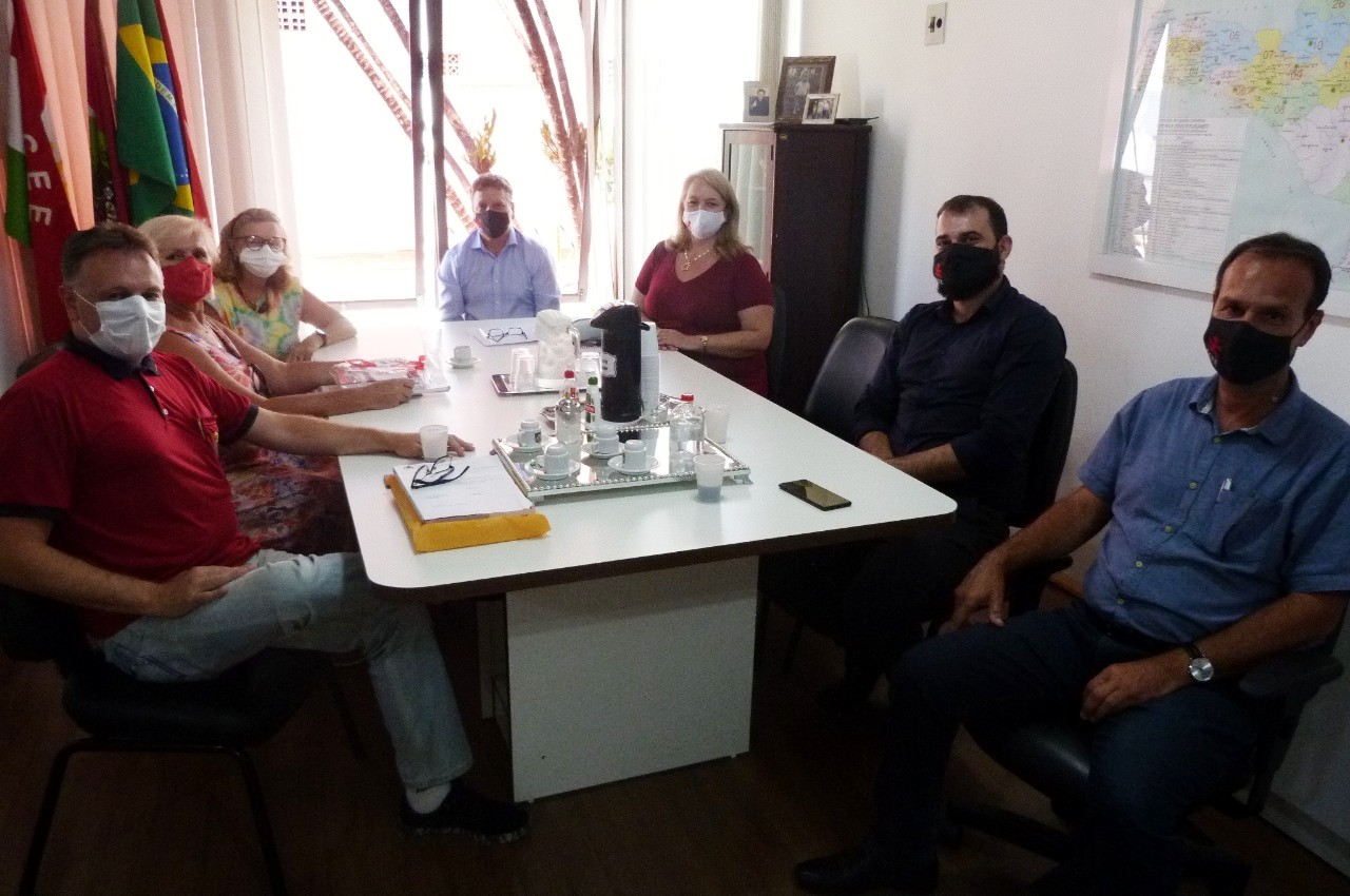 Sete pessoas sentadas em mesa de reunião posam para foto. Ao fundo bandeira do Estado. 