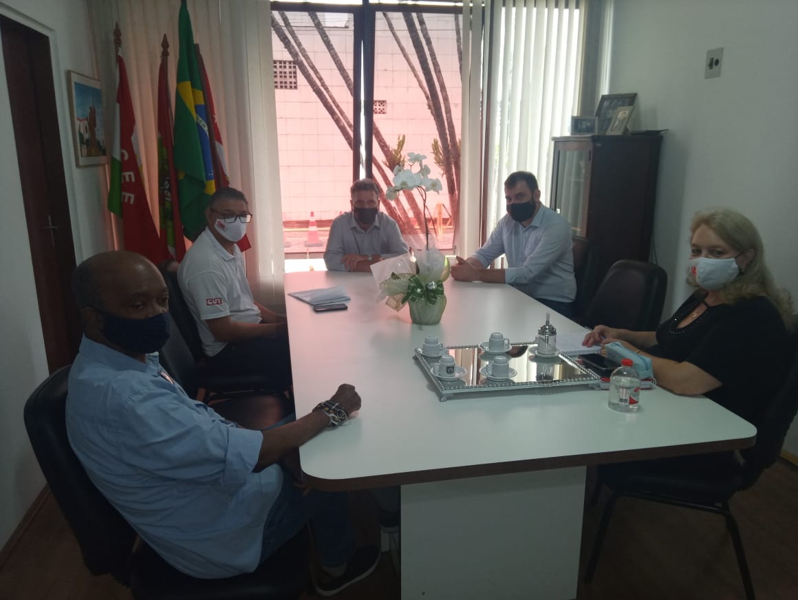 Cinco pessoas sentadas em mesa de reunião, ao fundo janela e bandeiras. 