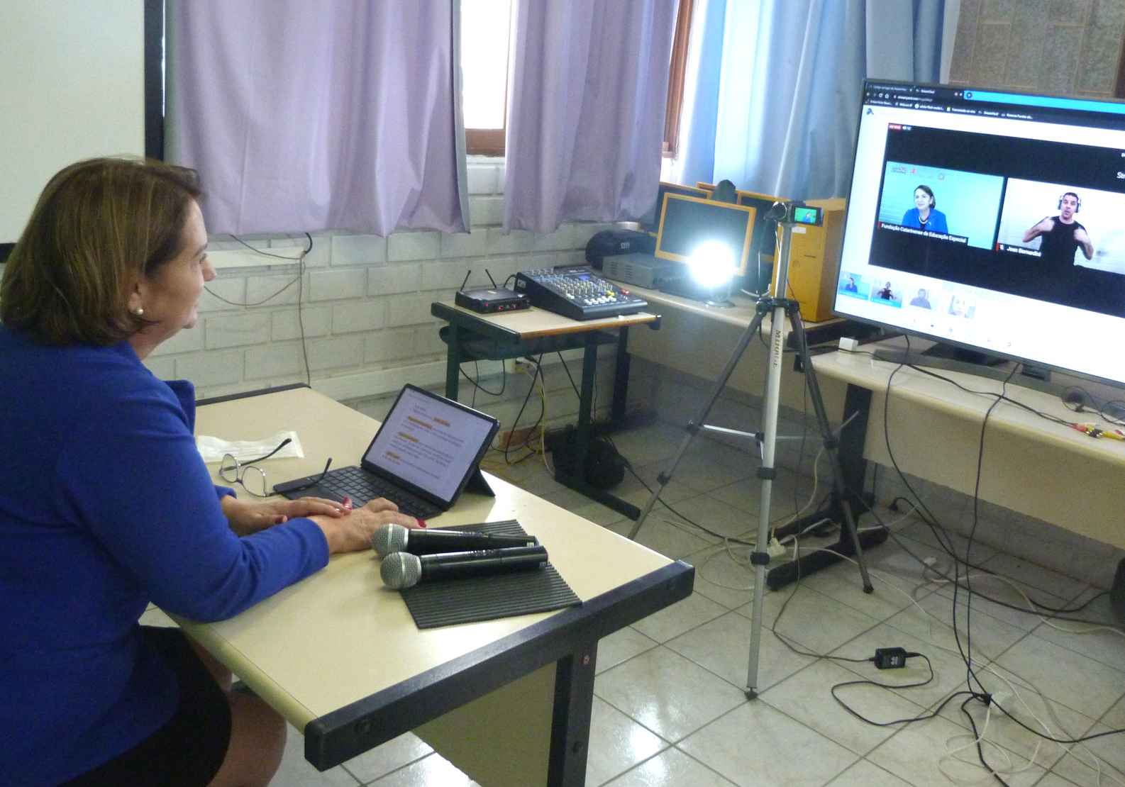 Mulher com terno azul sentada em frente a câmera e computadores 