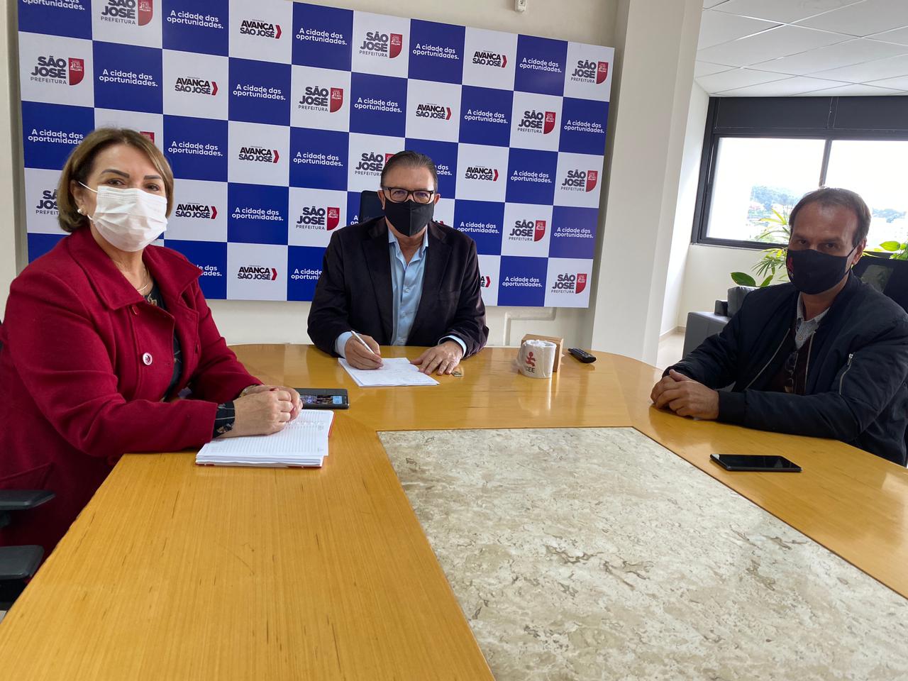 ambiente interno, 3 pessoas em mesa de reunião, uma mulher cabelos lisos e pelo ombro, casaco vermelho, e dois homens