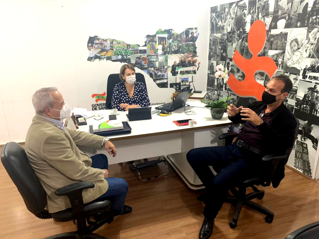 Sala interna, 3 pessoas sentadas conversam em torno de mesa