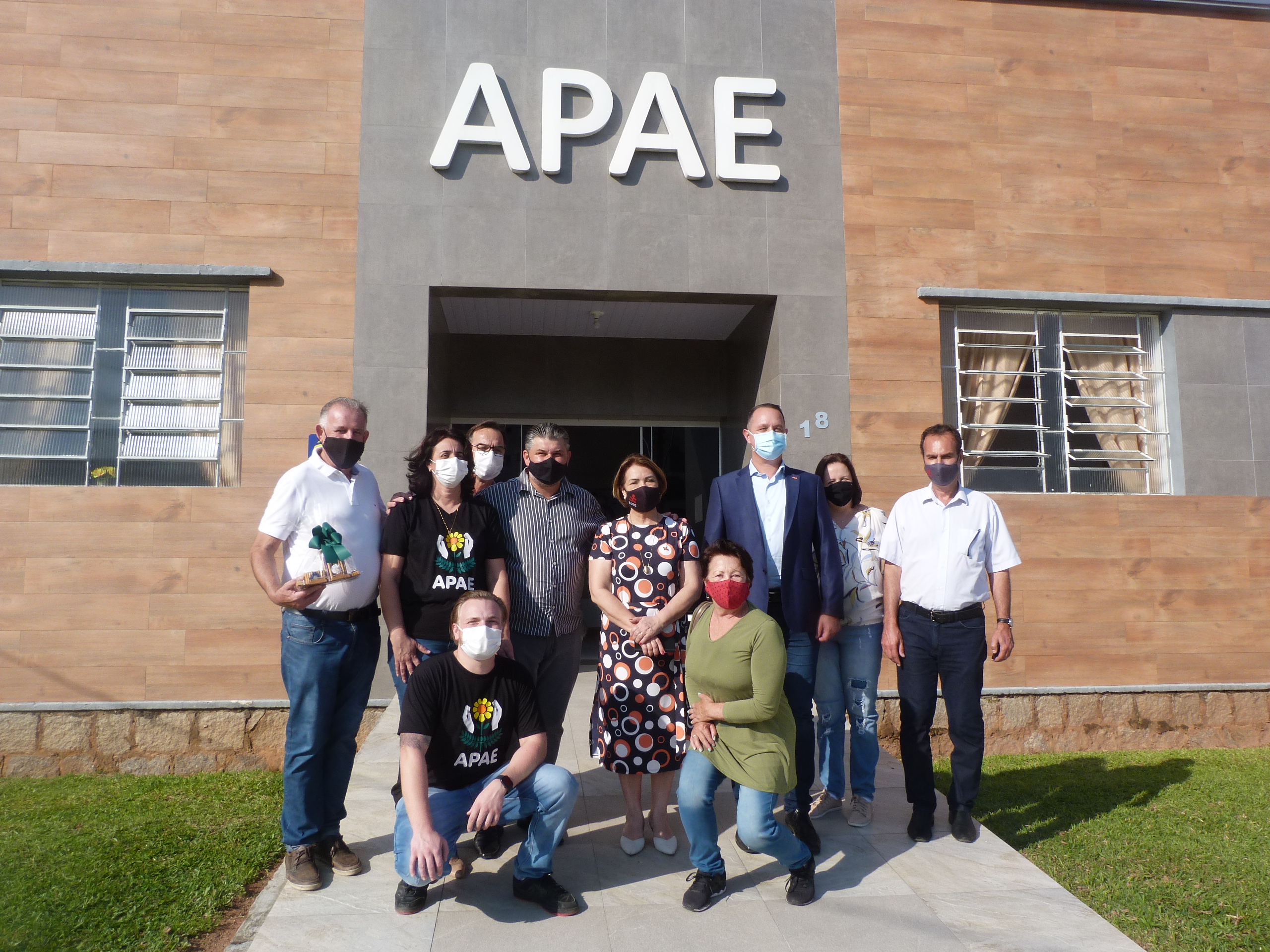 Oito pessoas em pé, posando para foto, ambiente externo, ao fundo prédio com letreiro Apae