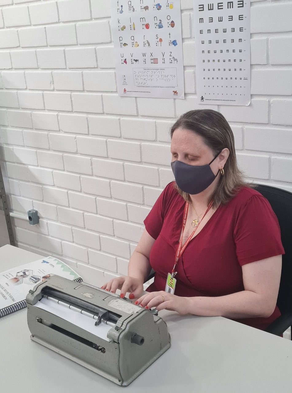 Foto de Josiane Becker escrevendo com a máquina braile. Ela está em uma sala com paredes de tijolinho brancas, sentada, com a máquina em cima de uma mesa de cor branca acinzentada. Josi é uma mulher branca, de cabelos castanhos com luzes na altura do ombro. Ela tem deficiência visual e está com os dois olhos fechados. Usa blusa com decote em V na cor vermelho escuro, unhas pintadas na mesma cor, brincos pequenos de argola dourada, colar com pingente dourado, máscara preta e crachá de identificação da FCEE. 