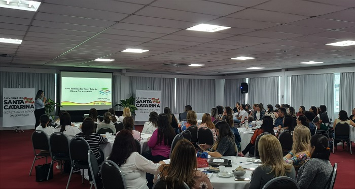 Foto de um auditório com várias mesas redondas e pessoas sentadas no entorno. O público assiste a uma aula. A professora aparece no fundo da imagem, em pé, ao lado do telão de slides. 