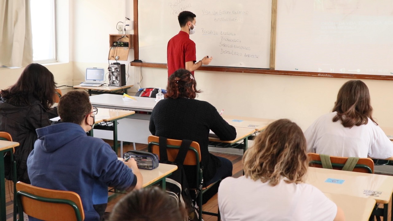 foto de uma sala de aula com 5 alunos e 1 professor. Todos estão de costas, olhando para o quadro, no qual o professor está escrevendo.