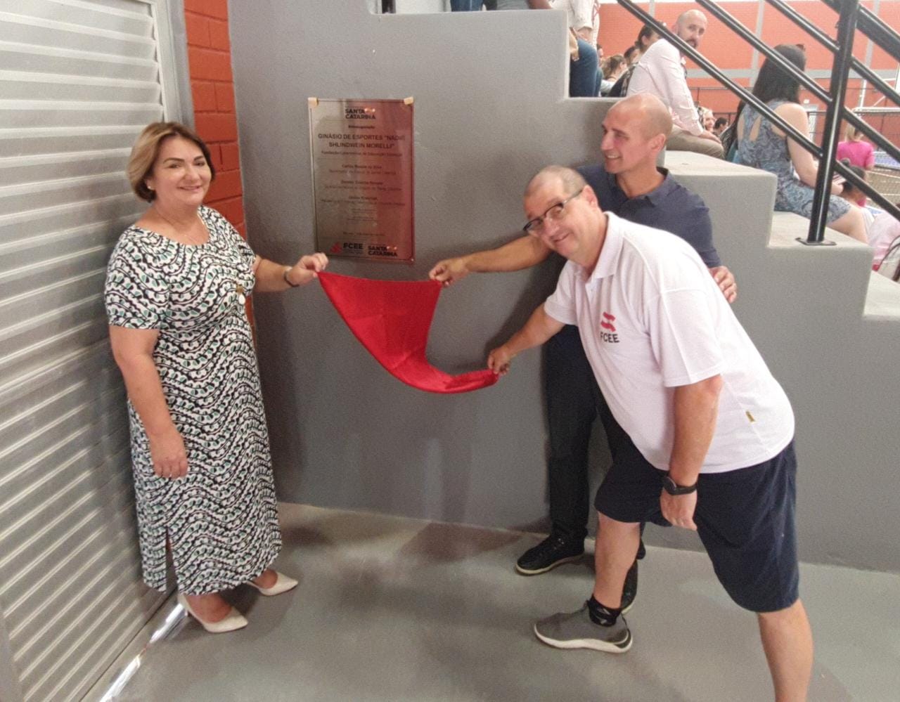 Ambiente interno, 3 pessoas puxam pano vermelho sobre placa de inox e posam para foto