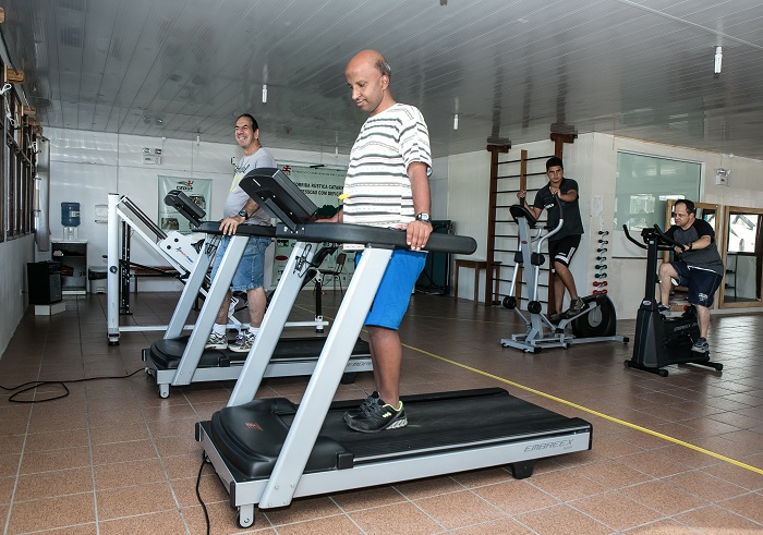 Foto de uma academia com esteiras e alguns outros aparelhos ao fundo. Em foco, dois homens estão praticando a esteira, concentrados. 