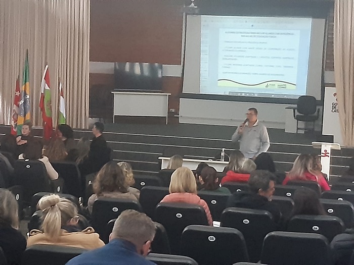 Foto de um auditório com um telão ao fundo transmitindo um slide e várias pessoas sentadas nas cadeiras